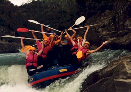 Six people in a rubber raft, wearing red life vests and yellow helmets pulling faces as they are white water rafting.